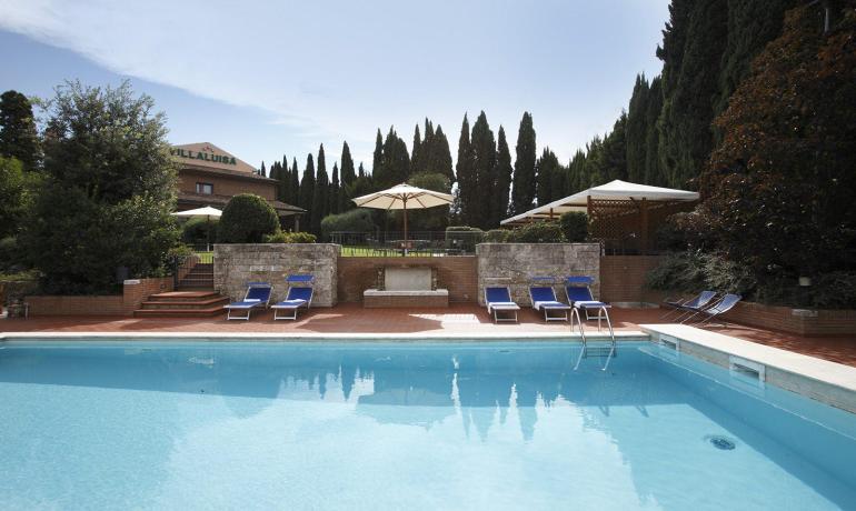 Piscina tranquilla circondata da cipressi e lettini blu sotto il sole.